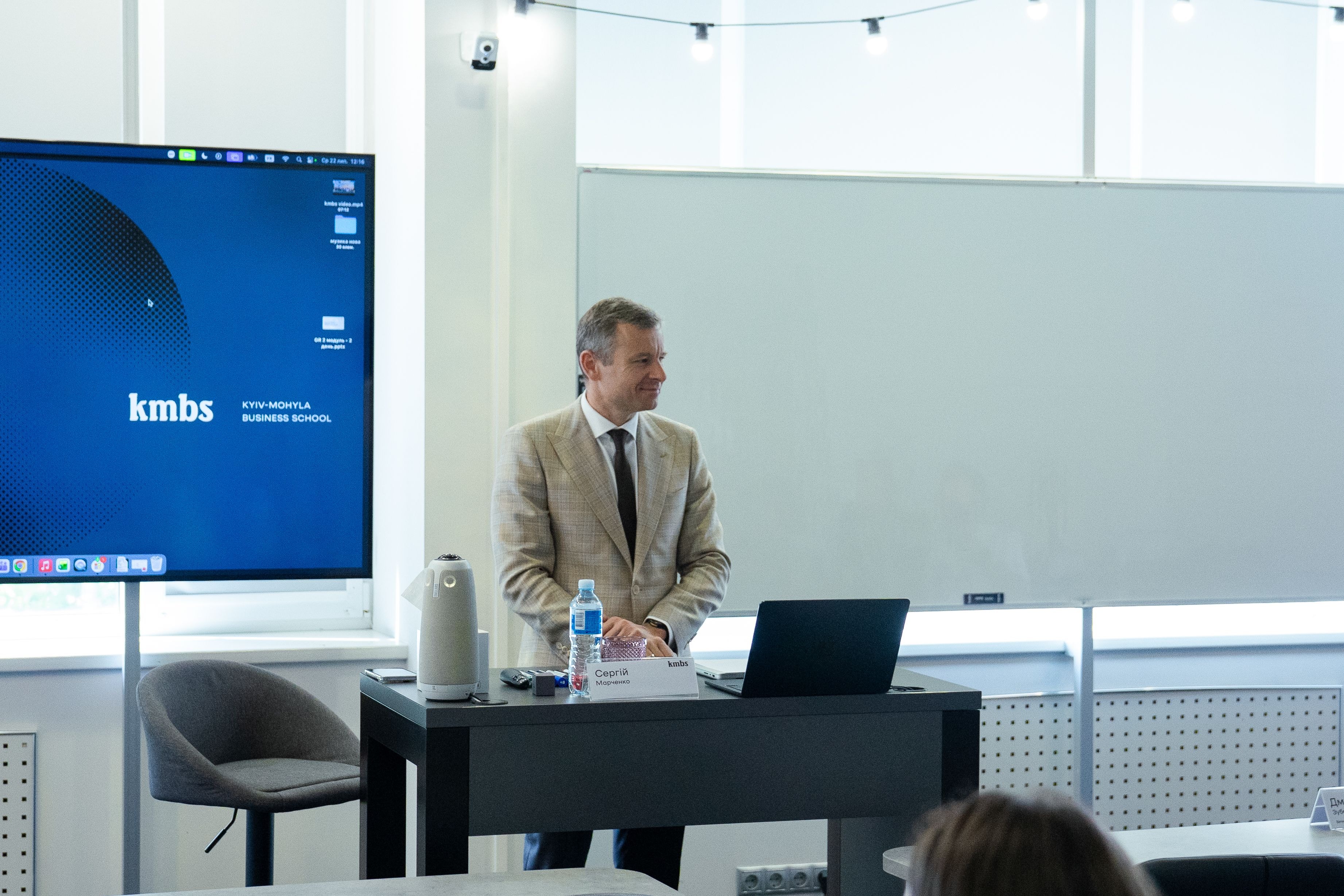 A man in a suit presents in a lecture hall with a large screen displaying "kmbs".