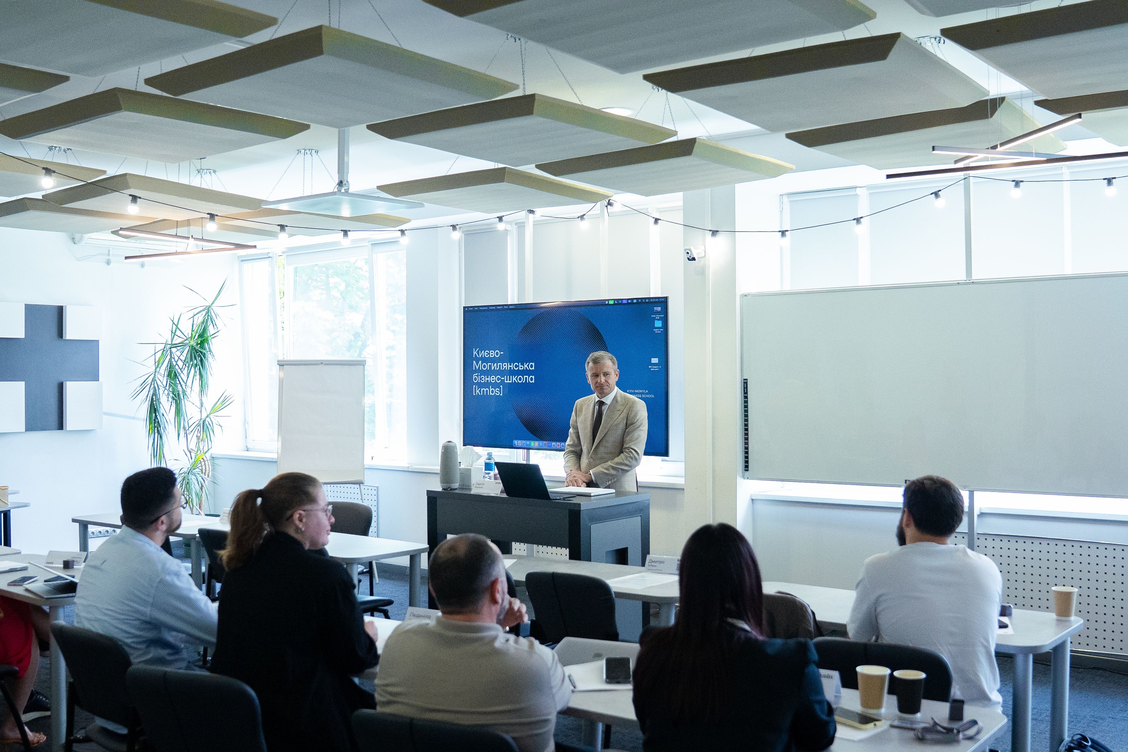 A speaker in a suit presents to an engaged audience in a well-lit modern conference room.