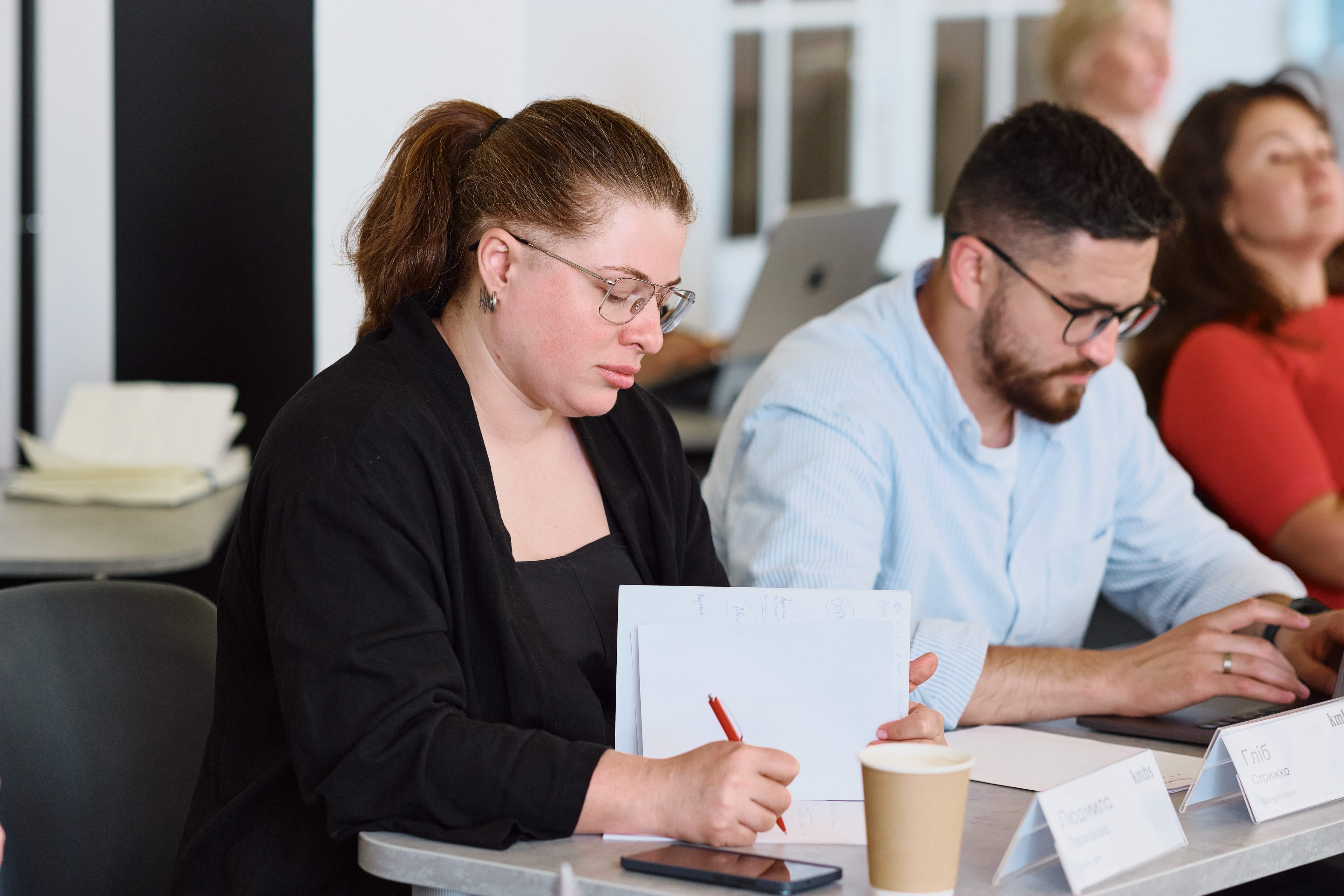 A woman in glasses focused on writing in a notebook during a learning session.