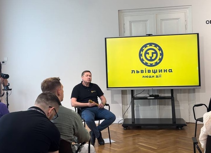 Man speaks at a presentation with a yellow 'Zusammenhalt' logo on a large screen.