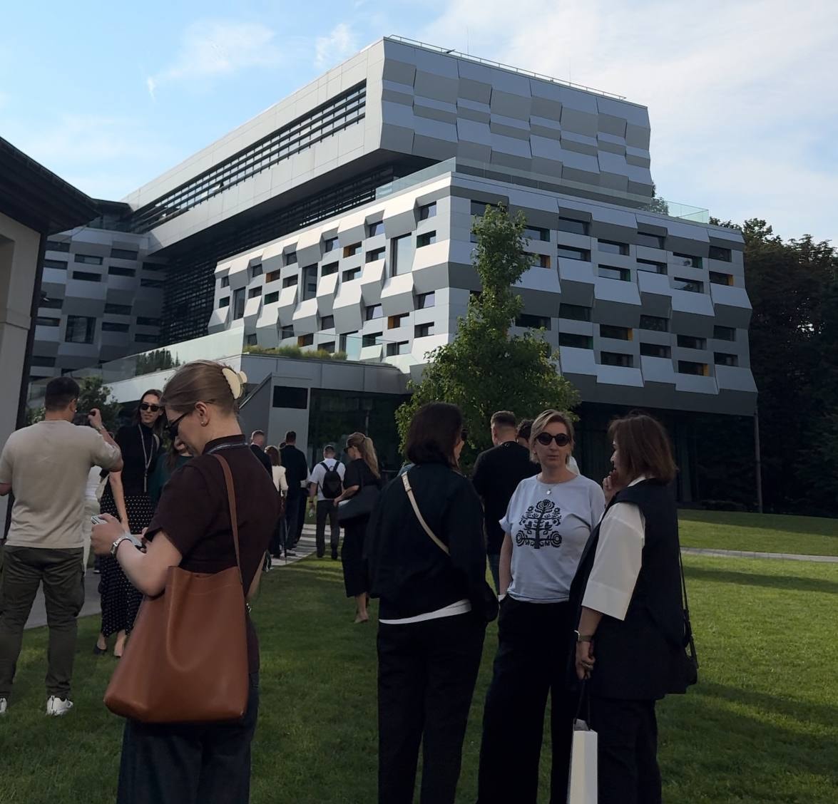 People gather on a grassy lawn in front of a striking modern architectural building.