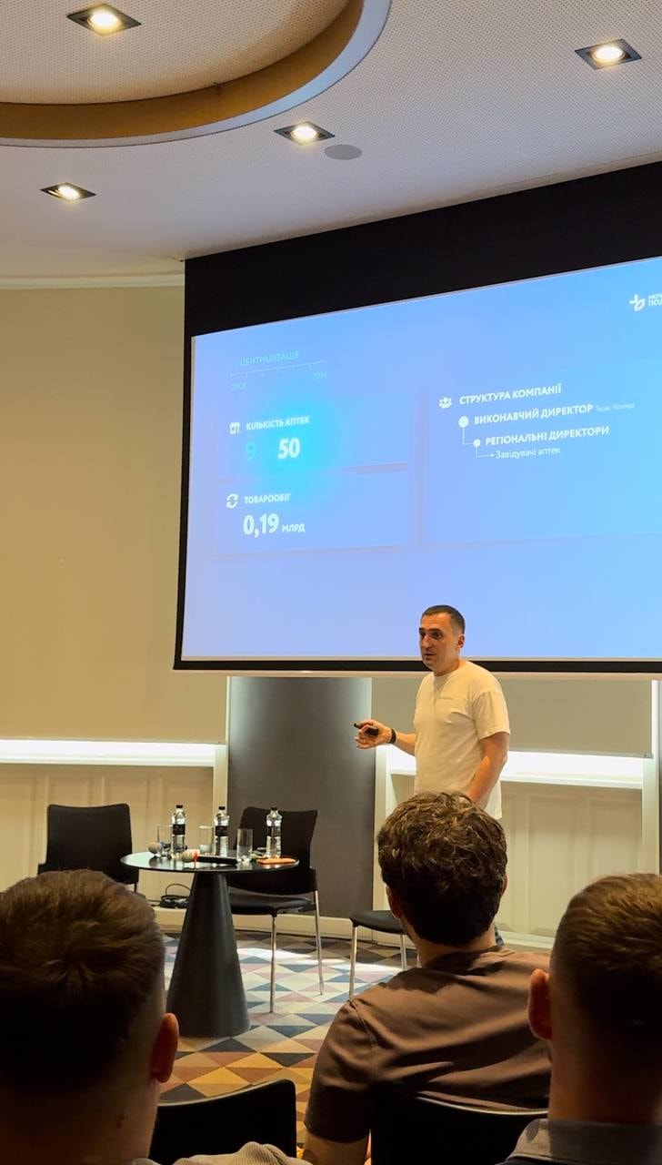 A man presents information on a large screen to an audience in a conference room.